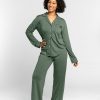 A person stands confidently, smiling, in a Harper FeatherTouch® Pajama Set by Woolx, featuring a temperature-regulating fabric, buttons, and a pocket on the shirt in Duck Green. The background is a plain light gray.