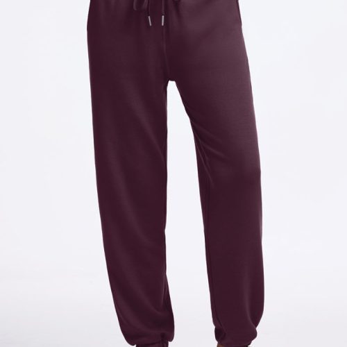 A person is wearing Woolx's Parker Sweatpants in Wine Tasting color with white sneakers, standing against a plain white background. These sweatpants provide travel comfort with features like a drawstring waist, elastic cuffs, and moisture-wicking properties to keep you cozy on the go. | Keesha is 5’9.5”, wearing a size S