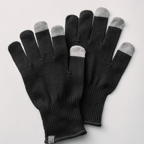 A pair of Asher Tech Gloves - Black by Woolx, featuring gray fingertips for touchscreen compatibility, is displayed on a light gray background. The gloves have ribbed cuffs and include a small tag on one glove.