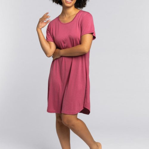 A person with curly hair smiles, wearing a knee-length, short-sleeved red violet Desi Nightgown OLD by Woolx. The cozy sleepwear complements their relaxed demeanor as they stand barefoot against a plain gray background.