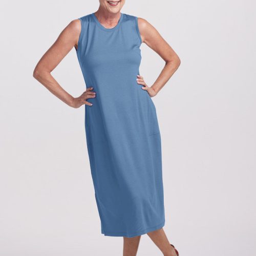 A woman with short white hair is smiling and posing in the sleeveless, knee-length Cassie Dress from Woolx, showcasing its lovely coronet blue color. She stands against a light background with one hand on her hip and the other resting by her side, complemented by brown sandals. The dress features practical side pockets for added charm, making it a perfect pick from the Stocking Stuffer Final Sale Deals.