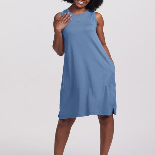 A person wearing the Woolx Clara Dress in Coronet Blue smiles with one hand touching their hair, standing casually with one leg slightly bent. They pair it with white sneakers and a necklace against a simple light gray backdrop.