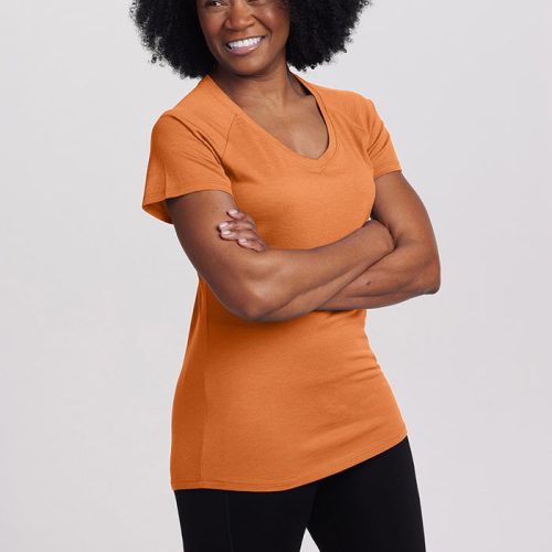 A person with curly hair smiles while standing with their arms crossed in a Woolx Mia Short Sleeve V Neck from the Stocking Stuffer Final Sale Deals! They are wearing orange and black pants against a plain background.