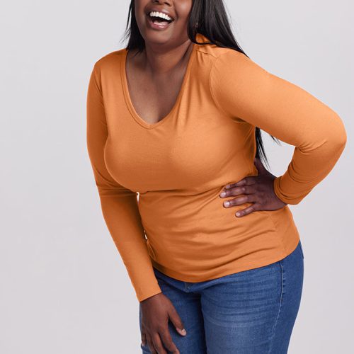 A person with long hair is delightedly smiling and laughing while wearing the Layla V Neck Top in orange from Woolx and blue jeans, standing against a plain background.