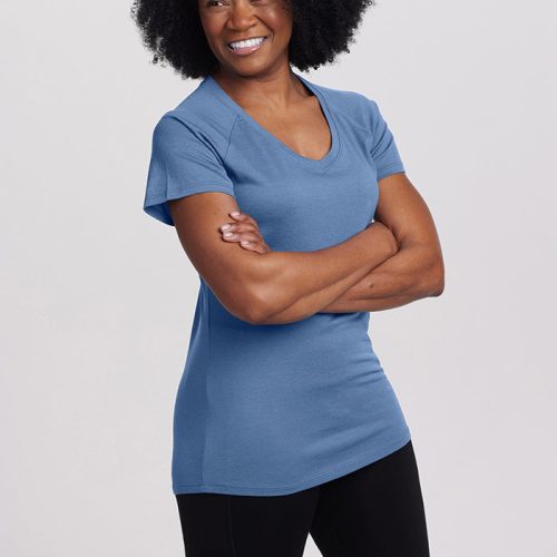 A person with curly hair smiles while standing with arms crossed, wearing a coronet blue Mia Short Sleeve V Neck from Woolx and black pants against a plain white background.