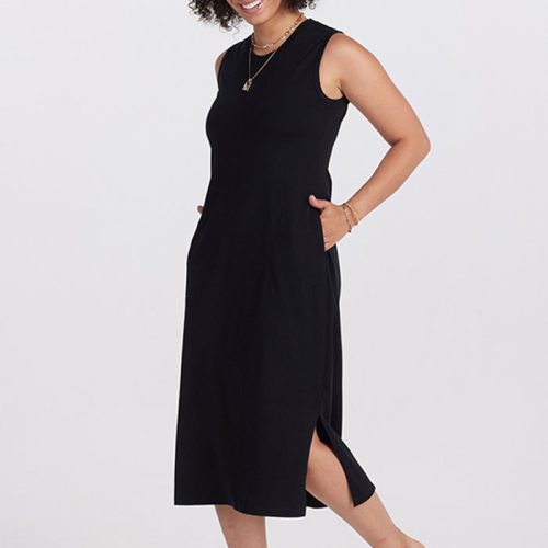 A person with curly hair smiles while wearing the sleeveless black Cassie Dress OLD from Woolx, paired with brown sandals, against a plain background. The breathable dress features a side slit, and they have their hands in pockets, accessorized with bracelets and a necklace.