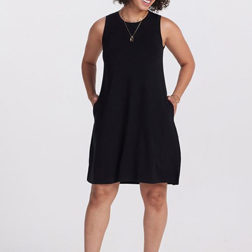 A person with curly hair stands smiling against a plain, light background, wearing a sleeveless black Woolx Clara Dress OLD and sandals. The stylish dress features pockets where they casually rest their hands. Completing the look is a necklace that adds a touch of elegance.