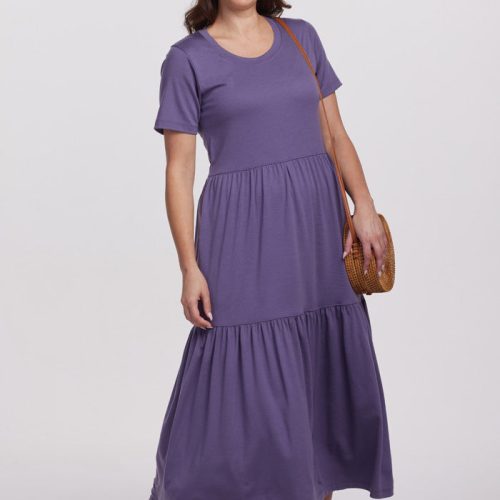 A woman in a stylish Montana Grape Lucia Dress by Woolx, paired with white sneakers, poses with a smile. The temperature-regulating merino wool fabric keeps her comfortable as she carries a round straw bag on her shoulder. The background is plain white.