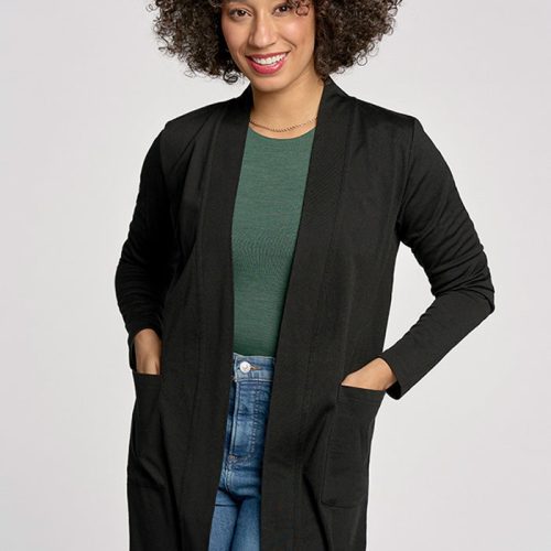 A person with curly hair is smiling and standing against a light background. They are wearing a green top, the black Paisley Cardigan by Woolx made of Australian Merino Wool, and blue jeans. Their hands are in the cardigan pockets.