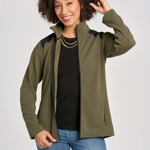 A smiling individual with curly hair is wearing a Woolx Peri Full Zip Jac in Olive over a black shirt and blue jeans, posing with one hand touching their collar. This Merino Wool piece is the perfect addition for any cold-weather wardrobe, set against a neutral background.