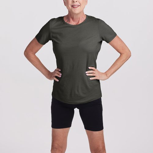 An older woman with short white hair stands confidently, hands on hips, wearing a lightweight Deep Fern Liza Crew Neck by Woolx and black shorts. She is smiling against a plain light gray background.