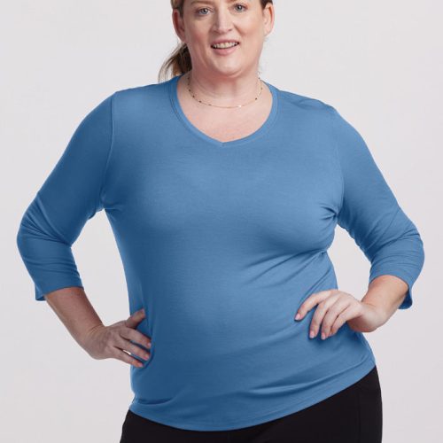 A person wearing a modern chic Elena VNeck in Coronet Blue by Woolx and black pants stands against a light grey background. They are smiling and have one hand on their hip.