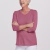 An elderly person with short white hair is smiling and standing with crossed arms, wearing a pink Elena VNeck from Woolx, part of the Stocking Stuffer Final Sale Deals, along with white pants crafted from relaxed fit Merino wool against a plain white background.