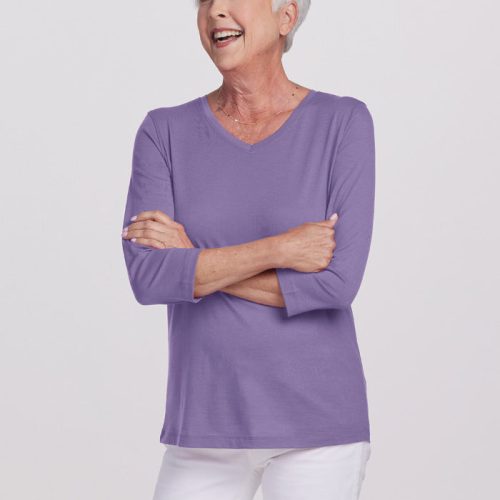 An elderly woman with short white hair smiles warmly, standing with her arms crossed. She's dressed in Woolx's relaxed fit Elena VNeck from the Stocking Stuffer Final Sale Deals, featuring three-quarter sleeves paired with white pants. The top's merino wool fabric ensures comfort against the plain white background.