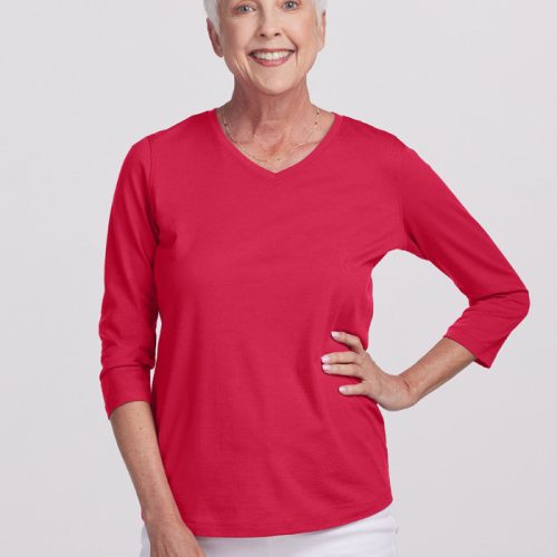 A mature individual with short white hair is beaming in a modern chic outfit, showcasing a Viva Magenta Elena V-Neck Top by Woolx paired with white pants. They stand confidently against a plain backdrop, one hand resting on their hip.