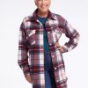 An older woman with short white hair smiles and poses confidently, wearing the Sutton Shacket in Frozen Cranberry Plaid by Woolx over blue jeans and a teal top. The Merino wool provides her breathable warmth as she stands against a plain white background, one hand on her hip.