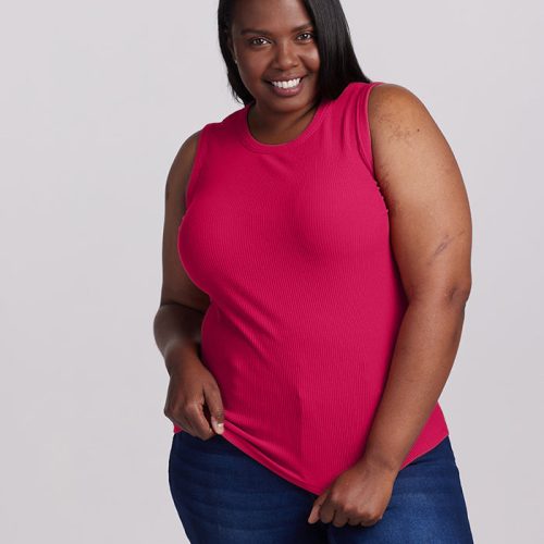 A person wearing a vibrant viva magenta Raegan Ribbed Tank Top by Woolx smiles confidently while posing. They have long dark hair and are shown against a plain gray background, with the ribbed texture of the tank harmonizing perfectly with their blue jeans.