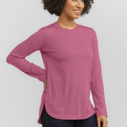 Smiling with curly hair and posing with one hand on their hip, a person showcases the relaxed fit of the Woolx Hazel Tunic, designed in a soft pink hue. They pair it with dark pants and stand against a light gray background.