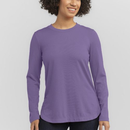 A person with curly hair is smiling, wearing a relaxed-fit, purple shirt reminiscent of the Hazel Tunic from Woolx and dark jeans, standing in front of a gray background.