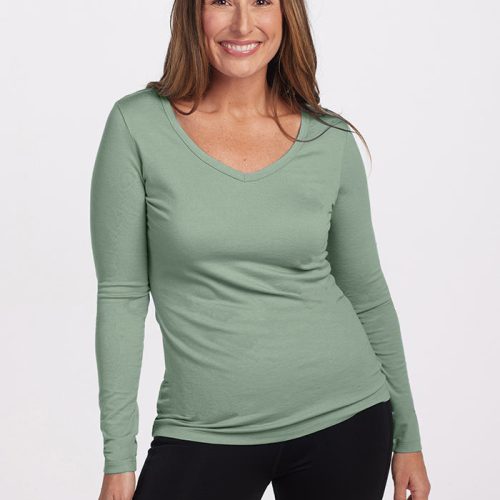 A person with long brown hair smiles while wearing the Woolx Layla V Neck Top in Basil, a long-sleeve shirt crafted from Australian Merino Wool, paired with black pants. The background is plain white.