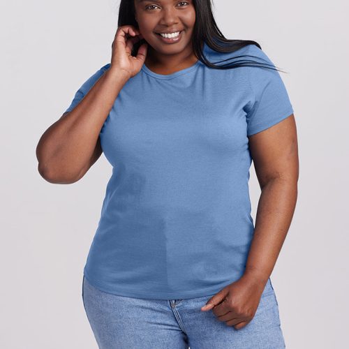 A smiling person with long hair is wearing the Woolx Liza Crew Neck in Coronet Blue and blue jeans, standing against a plain background. They have one hand on their hip and the other touching their neck.