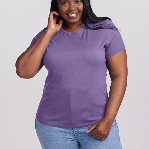 A woman with long dark hair smiles in a lightweight Montana Grape Liza Crew Neck tee and light blue jeans from Woolx. She holds one hand near her face, standing against a plain light gray background.