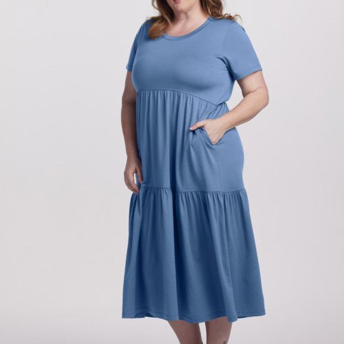 A woman wearing the Coronet Blue Lucia Dress from Woolx, featuring convenient pockets and crafted from temperature-regulating merino wool, stands against a plain background, gently smiling. She pairs the dress with brown sandals and has her hand placed in one of the pockets.