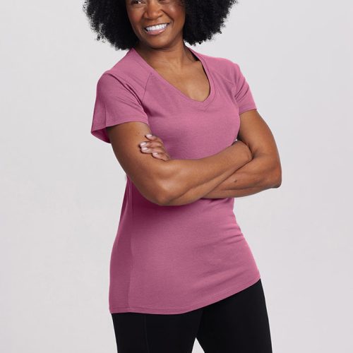 A person with curly black hair smiles while standing with their arms crossed, wearing a pink Mia Short Sleeve V Neck from Woolx and black pants against a plain light gray background.