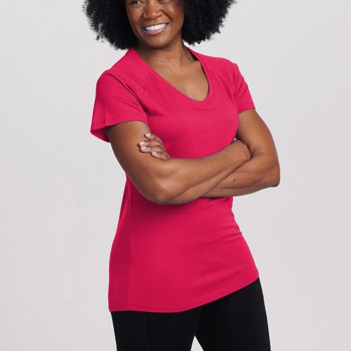 A person with curly hair smiles with arms crossed, wearing a Viva Magenta Mia Short Sleeve V Neck by Woolx and black pants, standing against a plain background. The ultra-soft material looks comfortable and stylish.