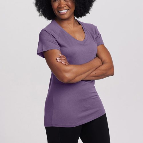 A person with curly hair smiles warmly, standing with arms crossed. They are wearing the ultra-soft Mia Short Sleeve V Neck in Montana Grape from Woolx and black pants. The background is plain and light-colored.