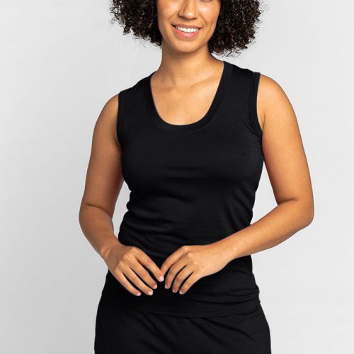 A person with curly hair smiles while standing against a plain background, wearing the Phoebe Lounge Tank in black by Woolx and matching shorts, embodying the essence of travel-friendly clothing. | Tori is 5'7", wearing a size S