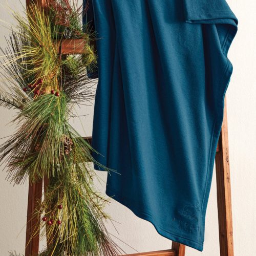 The Placid Merino Blanket by Woolx, in a real teal shade, is elegantly draped over a wooden ladder adorned with a festive garland of pine branches and red berries. This arrangement creates a warm, holiday-themed atmosphere against a light-colored wall.