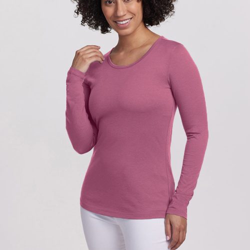 A person with curly hair is smiling, wearing a lightweight pink Remi Long Sleeve TShirt from Woolx, paired with white pants, and posing against a plain background.