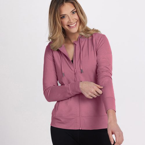 A person with long hair is wearing a Woolx Ryann Hoodie from the Stocking Stuffer Final Sale Deals in pink, paired with black pants, and standing against a plain white background. The lightweight hoodie enhances their cheerful expression as they are slightly turned to one side.