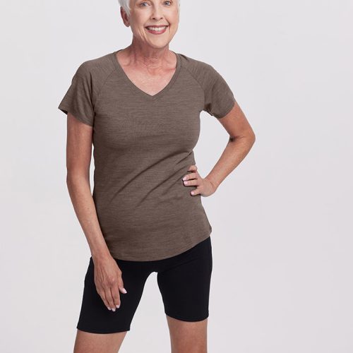 An elderly woman with short white hair confidently smiles in a Woolx Mia Short Sleeve V Neck and black shorts. She stands against a plain white background, her hand resting on her hip—a perfect look for any adventure, seamlessly combining style and comfort.