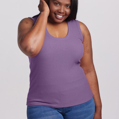 A person in a versatile fit, wearing a Montana Grape sleeveless Sloane Ribbed Tank by Woolx and blue jeans, smiles at the camera with one hand touching their ear against a plain, light-colored background.