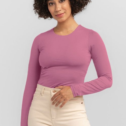 A person with curly hair is wearing a stylish long-sleeved top, likely the Sophia Bodysuit by Woolx in Mesa Rose, paired with beige pants. They are smiling and standing against a neutral background.