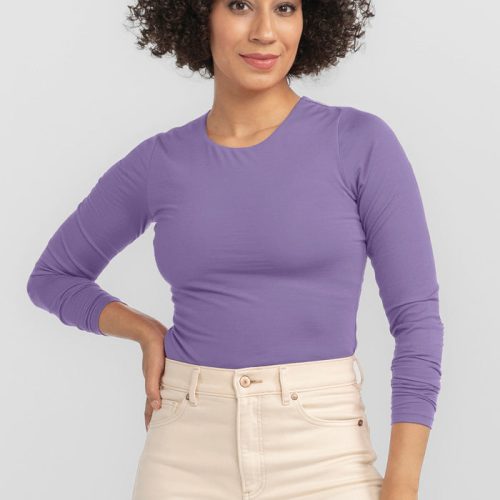 A person with curly hair is smiling, exuding a chic look in a long-sleeved Montana Grape Sophia Bodysuit from Woolx paired with light beige pants. They have one hand on their hip, and the background is plain light gray.