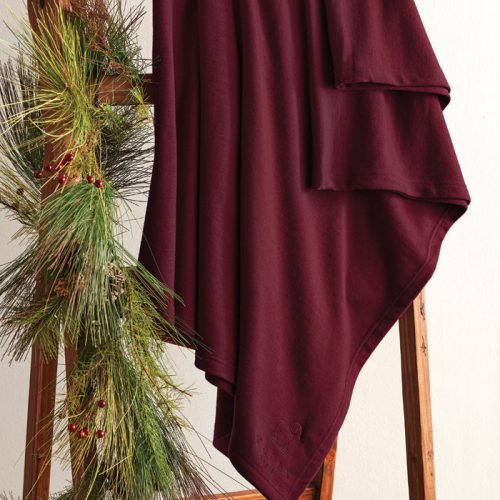 A Placid Merino Blanket in Windsor Wine, crafted by Woolx, is elegantly draped over a wooden ladder adorned with pine branches and red berries, creating a cozy and festive display against a light-colored wall.