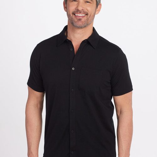 A bearded man with short, dark hair smiles as he stands against a plain white background, wearing the versatile black Endi Full Button Down Shirt from Woolx and blue jeans, certified by Woolmark®. | Matthew is 6', wearing a size L