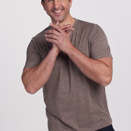 A man with short dark hair, wearing a Simply Taupe Endurance Tee by Woolx and dark jeans, smiles and clasps his hands against a plain white background. | Brandon is 6’3.5”, wearing a size XL