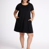 A woman is standing and smiling in the Georgie Dress - Black by Woolx, with her hands in the side pockets. She is wearing white sneakers and is posed against a plain white background. | Juliana is 5'10", wearing a size XL