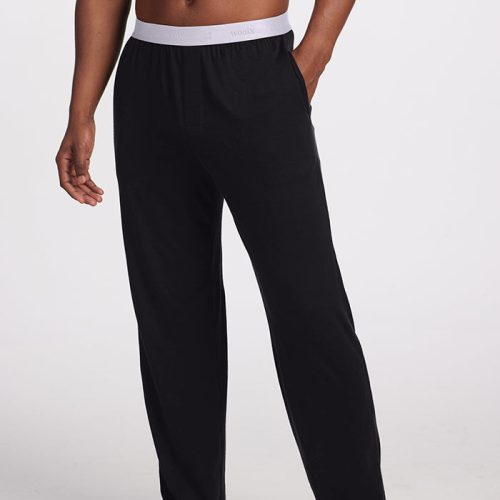 A person is standing barefoot on a white surface, wearing the Woolx Arlo Lounge Pants in black. These temperature-regulating Merino wool pants feature a white waistband and are designed with a long, loose fit, making them ideal for casual wear or sleepwear. The background is simple and white.