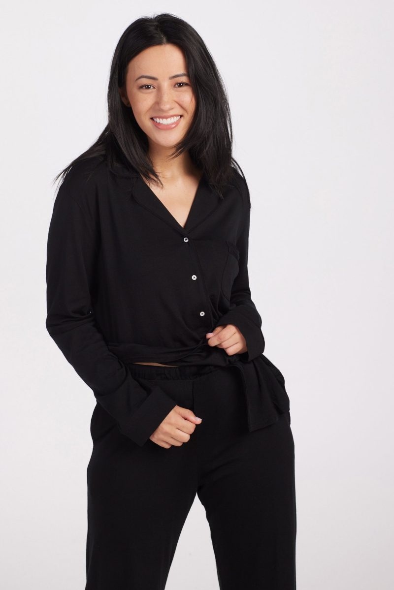 A person with long black hair smiles while standing in a relaxed pose, effortlessly stylish in the Harper FeatherTouch® Pajama Set by Woolx, featuring a black long-sleeve button-up top and matching pants made from temperature-regulating merino wool, against a plain white background.