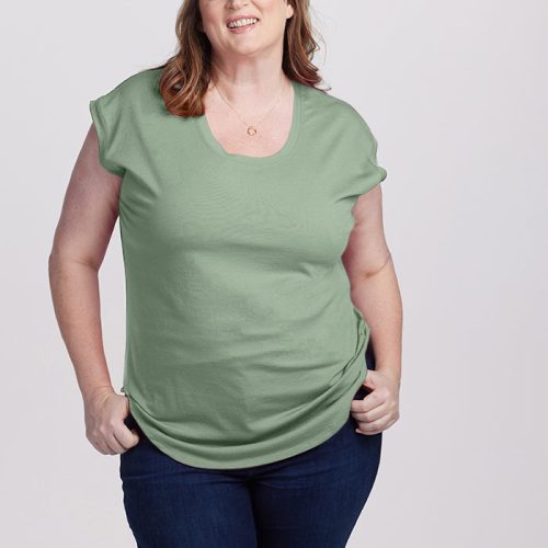 A person with shoulder-length brown hair is smiling and wearing the Gabi Tunic Top in Basil by Woolx, paired with dark jeans. Their hands are in their jeans pockets as they stand against a plain, light-colored background.