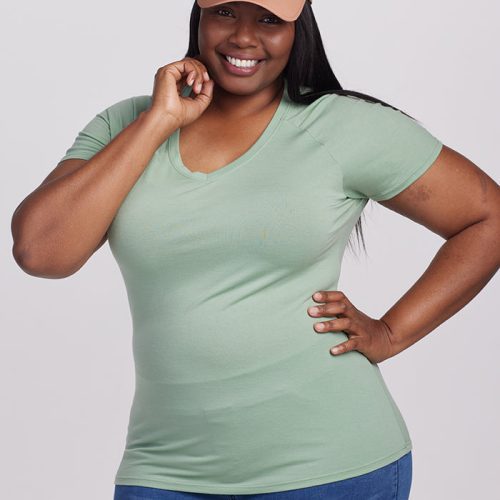 Dressed in an ultra-soft Mia Short Sleeve V Neck in Basil by Woolx, a person smiles while touching their face with one hand. Their other hand rests on their hip against the plain white background, complementing their blue jeans and beige cap.