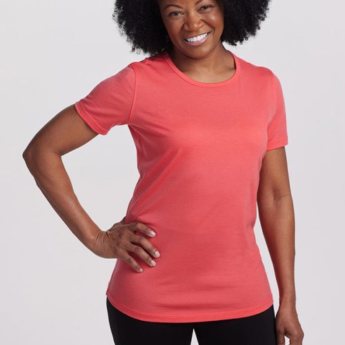 A person with curly black hair is smiling and wearing a lightweight Cayenne-colored Liza Crew Neck tee from Woolx, along with black pants, standing against a plain light background.