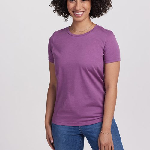 A person with curly hair smiles while wearing a Dewberry Liza Crew Neck by Woolx and blue jeans, standing against a plain white background.