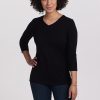 A person with curly hair smiles, exuding a casual chic style while standing with one hand on their hip. They are wearing the sleek black Elena V Neck from Woolx paired with dark jeans, set against a plain white background.