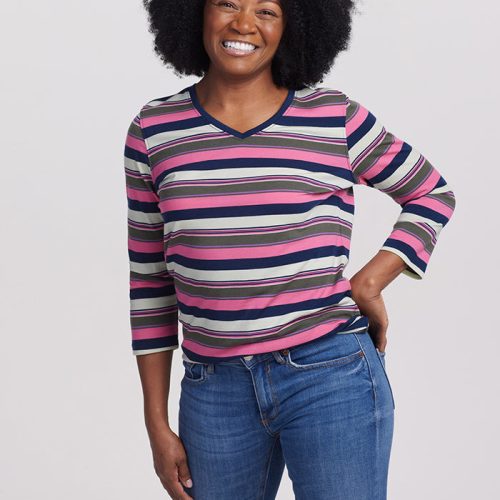 A person with curly black hair beams warmly while wearing a modern and chic Elena VNeck Top from Woolx, featuring Aurora Stripe colors of purple, pink, and gray. Their left arm is bent and rests on their hip as they pair it with blue jeans against a plain white background.